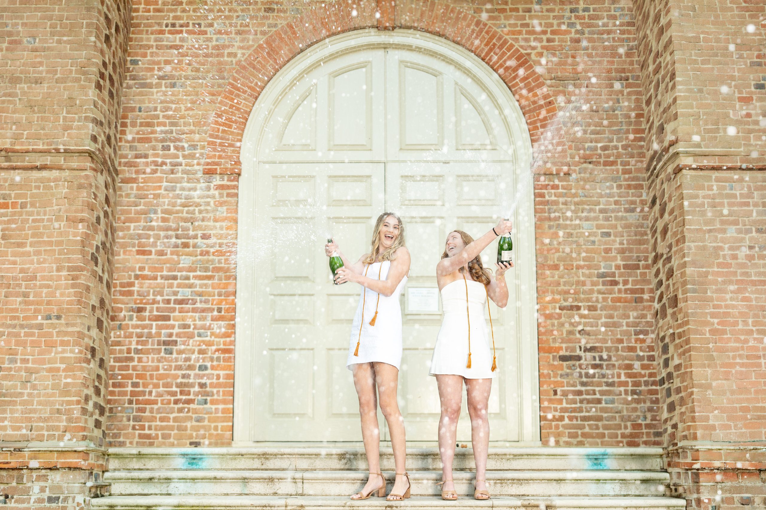 Two graduates in white dresses popping champagne at an arched brick doorway