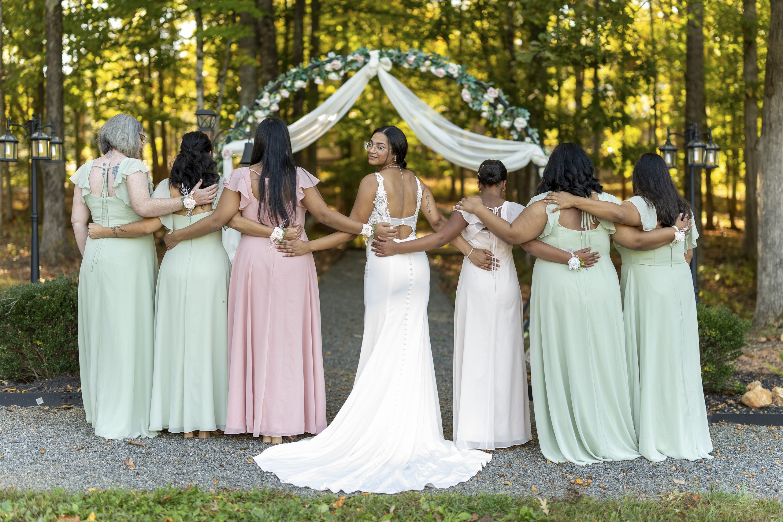 Bride with her bridesmaids in sage and pink gowns beneath a floral ceremony arch